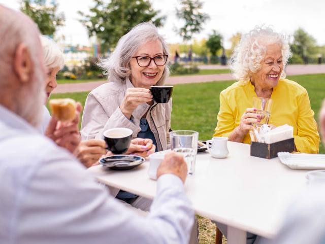 Older adults sharing coffee at an outdoor cafe in a park, smiling and chatting.