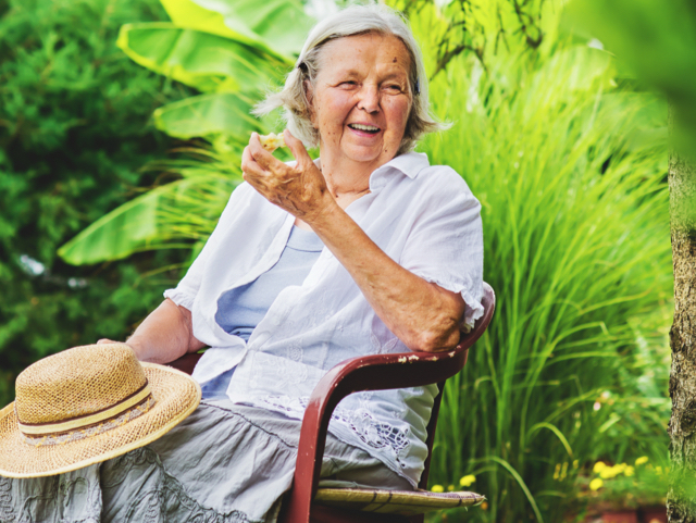 Older woman sitting on a chair in a garden, smiling with a straw hat resting on her lap